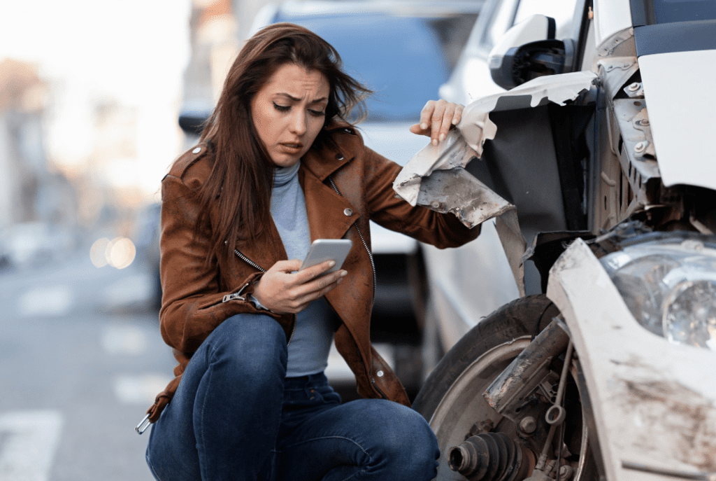 A worried woman looks at her phone while examining extensive damage to her car